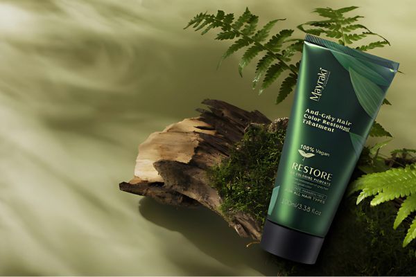 A green tube of Mayraki Anti-Gray Hair Color Restoring Treatment rests on a mossy log surrounded by ferns, emphasizing a natural, botanical aesthetic. The label promotes the product as 100% vegan and designed to restore hair’s natural pigment.
