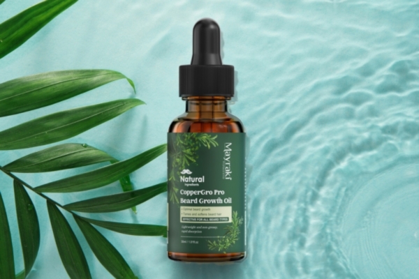A dropper bottle of Mayraki CopperGro Pro Beard Growth Oil placed on a light blue water ripple background with tropical green leaves on the side, emphasizing a fresh and natural look.
