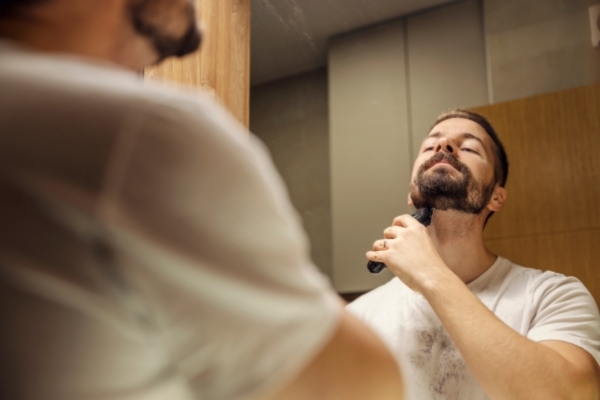 A man trims his beard using an electric shaver while looking into a bathroom mirror, with small hair clippings visible on his white t-shirt.