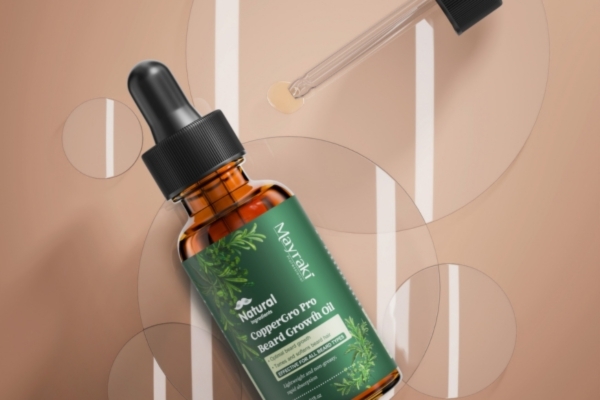 A brown glass dropper bottle labeled "Mayzak Natural CopperGro Pro Beard Growth Oil" lies on a warm-toned surface with overlapping translucent circles. A dropper rests nearby with a droplet of oil at the tip, emphasizing the product's natural ingredients and premium packaging.