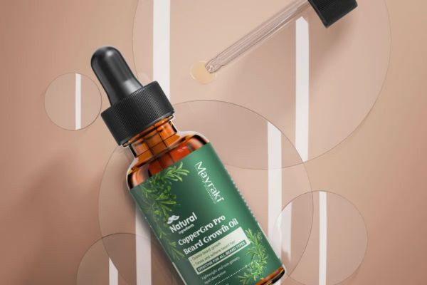 Mayraki Natural CopperGro Pro Beard Growth Oil bottle lies on a soft beige background with a dropper placed above it, dispensing a small amount of oil. The layout highlights product texture and packaging design.