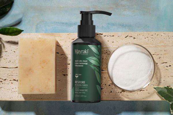 A bottle of Mayraki Anti-Gray Hair Color Restoring Treatment sits on a stone surface between a natural bar of soap and a dish of white powder. The bottle features green botanical graphics, emphasizing its vegan, paraben-free, and sulfate-free formula.