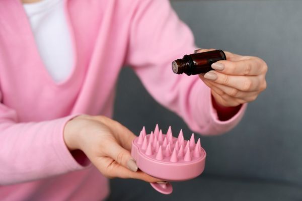 Close-up of a person in a pink sweater pouring essential oil from a small brown bottle onto a pink silicone scalp massager with soft spikes. The setting suggests a self-care or haircare routine focused on enhancing scalp massage benefits.