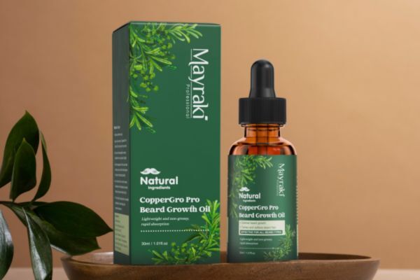 A bottle of Mayraki CopperGro Pro Beard Growth Oil is displayed next to its matching green box, both labeled with natural ingredients like ginseng and aloe vera. The setup is styled with a wooden bowl and greenery, emphasizing a clean, botanical aesthetic.