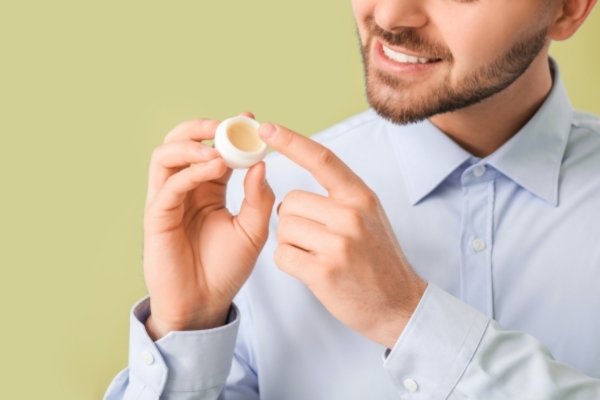 A smiling man in a light blue button-up shirt holds a small open jar of grooming balm and scoops some with his finger. The image reflects a clean and confident grooming routine, complementing Hair Mayraki’s dedication to premium beard and skincare solutions.
