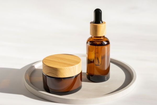 A brown glass jar with a wooden lid and a matching amber dropper bottle sit on a round ceramic tray, bathed in soft natural light. The minimalist setup highlights the contrast between beard balm and beard oil in stylish, eco-conscious packaging.