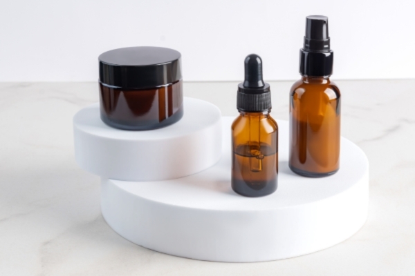 Beard Butter vs Beard Oil vs Beard Balm Three amber-colored cosmetic containers—a jar, a dropper bottle, and a spray bottle—are arranged on sleek white display pedestals against a neutral background. The setup emphasizes clean, minimalist skincare or grooming product presentation.