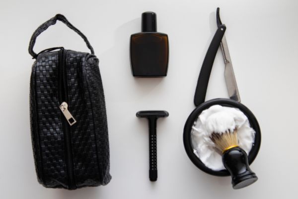 Flat lay of travel-friendly grooming tools including a black toiletry bag, a razor, shaving cream with a brush, a straight razor, and a small bottle of cologne, all neatly arranged on a white surface.