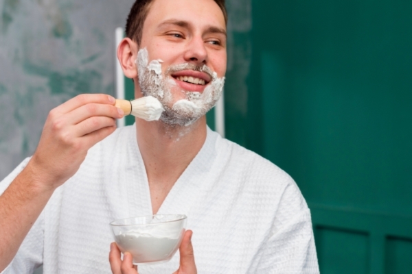 Smiling man in a white bathrobe applying shaving cream to his face with a brush, holding a bowl of cream in his other hand in a bathroom setting.