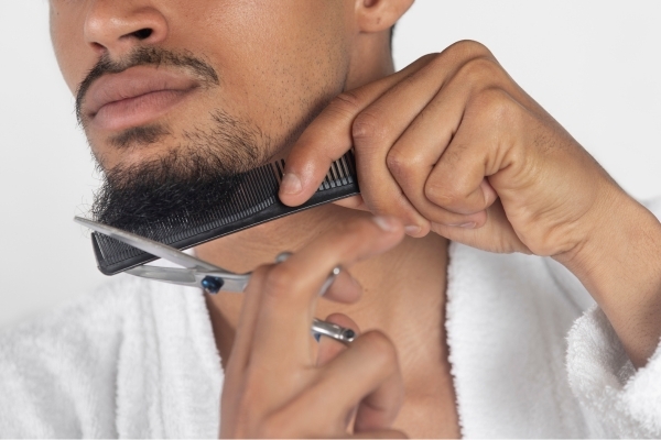 Beard Care Habits That Support Healthy Growth A man in a white robe carefully trimming his beard with scissors while using a comb to guide the shape, illustrating proper grooming as part of healthy Beard Care Habits.