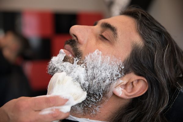 A man with long hair and a full beard relaxes with his eyes closed as a barber applies shaving cream to his face using a brush. The scene highlights grooming or beard care preparation, possibly in relation to maintaining a healthy beard in outdoor conditions.