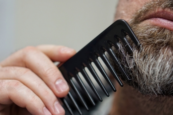 A close-up of a man's hand using a wide-tooth black comb to groom his gray-streaked beard, focusing on beard maintenance and care.