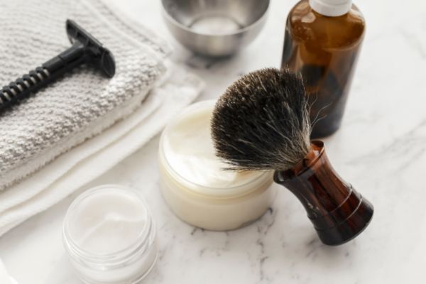 A classic shaving setup featuring a shaving brush resting on a jar of cream, a safety razor, a brown glass bottle, a towel, and a metal bowl arranged neatly on a marble countertop.