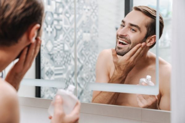 Smiling man applying product to his beard while looking in the mirror, holding a small bottle in one hand. The bright bathroom setting and his cheerful expression suggest a positive grooming routine, likely involving a beard-strengthening protein treatment.