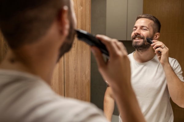 Man smiling in the mirror while grooming his beard with an electric trimmer, wearing a white t-shirt in a modern bathroom with wood paneling.