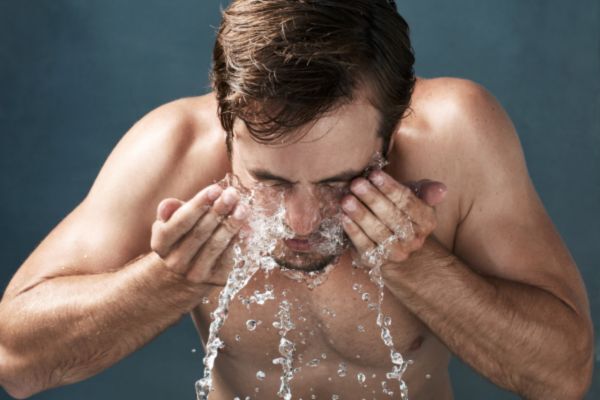Shirtless man splashing water on his face, mid-wash, with water droplets suspended in motion against a dark background, emphasizing a fresh and cleansing skincare routine.