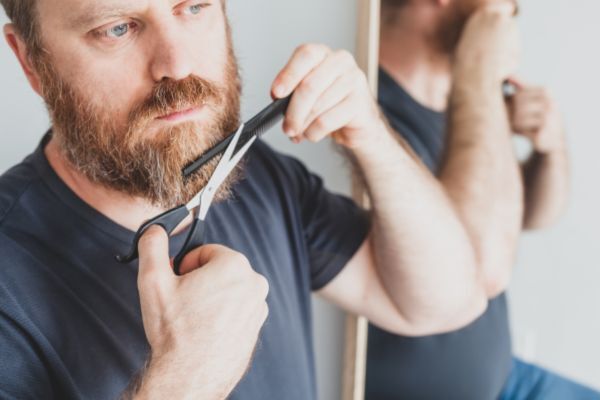 Alt text: A bearded man carefully trimming the ends of his beard with scissors while looking in a mirror, wearing a dark t-shirt and concentrating on grooming.