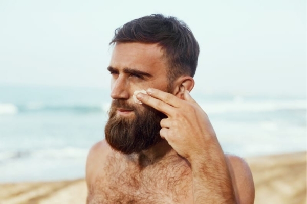A shirtless man with a full beard stands on the beach applying sunscreen to his cheek, illustrating Beard Sun Protection Tips for keeping facial hair and skin healthy under the sun. The image supports advice from Hair Mayraki on using SPF and natural oils for UV protection.