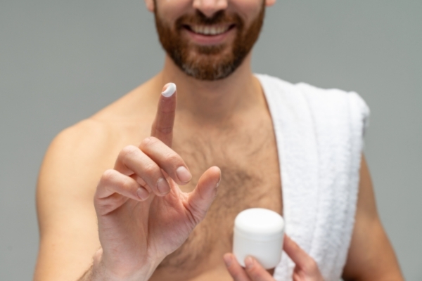 Smiling man with a trimmed beard holds a small white jar and shows a fingertip dab of cream, wearing a white towel over his shoulder. The image suggests a skincare or grooming routine, emphasizing personal care.