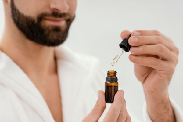 A man in a white bathrobe holds a small amber bottle of beard oil, using a dropper to extract a few drops. He has a neatly groomed beard and a slight smile, highlighting a grooming routine.