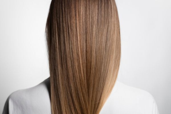 A back view of a person with long, straight, and silky light brown hair, appearing smooth and frizz-free, wearing a white top against a neutral background.