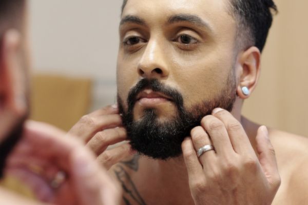  A man with a neatly groomed beard examines his facial hair closely in the mirror, gently touching his beard with both hands. He wears a silver ring and a small gauge earring, appearing focused on his grooming routine.