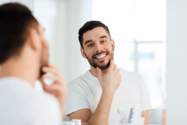 A man with a neatly groomed beard smiles while admiring his reflection in the bathroom mirror, gently touching his chin. He appears confident and satisfied, suggesting the benefits of using beard oil in his grooming routine.