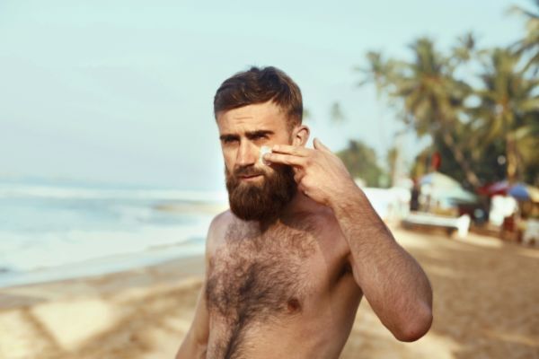 A shirtless man with a full beard stands on a tropical beach, applying a product to his face with two fingers. Palm trees, sand, and ocean waves create a relaxed, sunny backdrop, suggesting skincare or beard care in a beach setting.