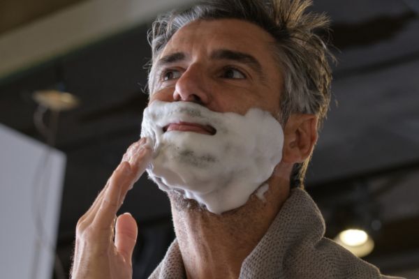 A man with salt-and-pepper hair applies a thick layer of shaving foam to his face while looking upward. He’s dressed in a bathrobe, suggesting a calm grooming routine at home.