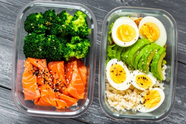 Two clear meal prep containers filled with vibrant, healthy ingredients on a dark wooden surface. One contains raw salmon and steamed broccoli, while the other includes sliced avocado, halved boiled eggs, and brown rice, all topped with black and white sesame seeds.