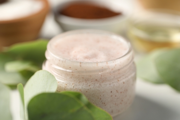 Open jar of natural exfoliating beard scrub with visible fine granules, surrounded by green leaves and oils, representing the benefits of beard scrub in removing buildup and promoting healthier skin beneath the beard.