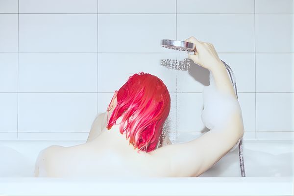 A person with vibrant pink hair sits in a bathtub, rinsing their hair under a handheld showerhead, surrounded by white tile. This image captures a gentle washing routine, reinforcing tips on how to protect colored hair from damage during cleansing.
