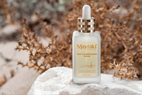A bottle of Hair Mayraki Hair Growth Intense Serum with a gold dropper cap is displayed on a white stone against a background of dried foliage. The elegant packaging and natural setting highlight Hair Mayraki’s targeted approach to promoting hair regrowth through science-backed ingredients.