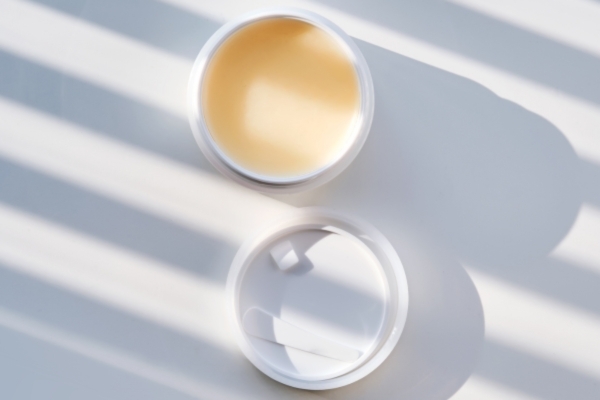 An open container of solid balm under striped light, representing a natural butter-based product used in beard care routines to lock in moisture and enhance softness.