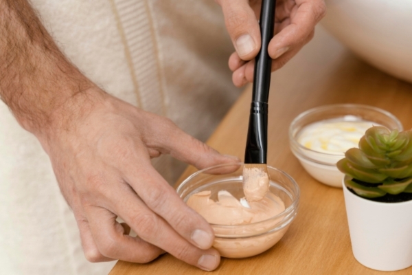 A person dips a cosmetic brush into a small glass bowl filled with a creamy pink facial mask, with another bowl of white cream and a small potted succulent nearby on a wooden surface.