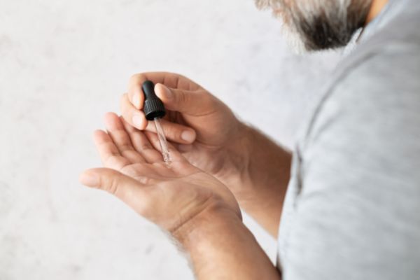 A man applies beard oil from a glass dropper onto the palm of his hand. His face is partially visible, showing a graying beard and a gray shirt.