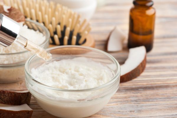 A glass bowl filled with a creamy, coconut-based hair treatment is surrounded by coconut slices, a dropper, a wooden hairbrush, and an amber bottle. The natural setup suggests DIY or organic protein treatments for hair nourishment and repair.