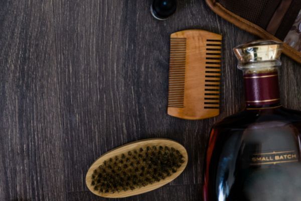 A flat lay of beard grooming essentials on a dark wood surface, including a wooden comb, a bristle beard brush, and a glass bottle with a maroon label.