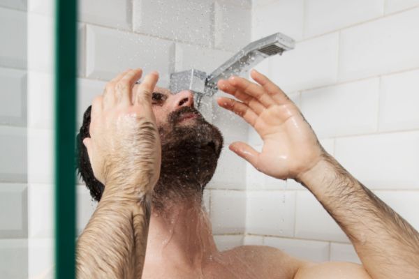 A man with a beard stands under a showerhead in a tiled bathroom, tilting his head back as water sprays onto his face. His hands are raised near his head, suggesting he's rinsing or adjusting to the water.