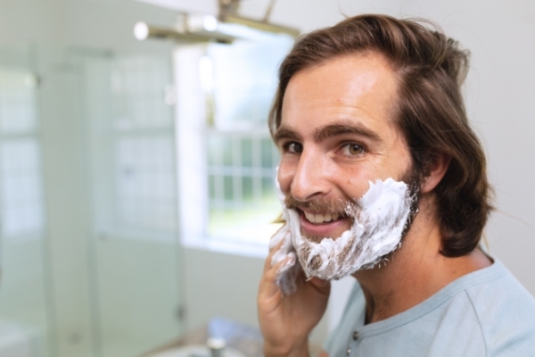 Cleansing / Washing Man smiling while applying shaving cream to his beard in front of a bathroom mirror, illustrating the role of regular beard grooming in maintaining facial hair cleanliness and shape.