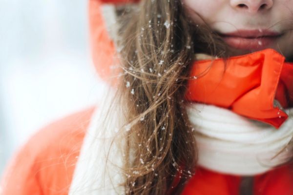 Close-up of a woman’s face and hair in snowy weather, with snowflakes caught in her long brown hair. She’s wearing a bright orange winter coat and a white scarf, highlighting the effects of cold weather on hair.