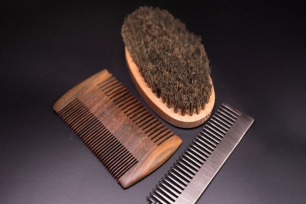 Flat lay of three beard grooming tools on a dark surface: a boar bristle brush with a wooden handle, a wooden comb, and a metal comb. The image highlights different textures and materials for effective beard maintenance and styling.