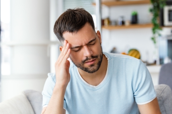 A man sitting indoors with his eyes closed and his hand pressed to his temple, appearing to be in pain or deep thought. He wears a light blue shirt and has a tense facial expression, suggesting a headache or stress.