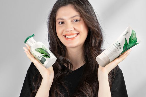 A smiling woman with long, dark hair holds up two Mayraki hair care products—“Ultra Repairing” and “Silky Smooth Frizz-Free Hair Treatment Shampoo”—in each hand. The white and green packaging features botanical designs, emphasizing a natural approach to hair repair and smoothing.