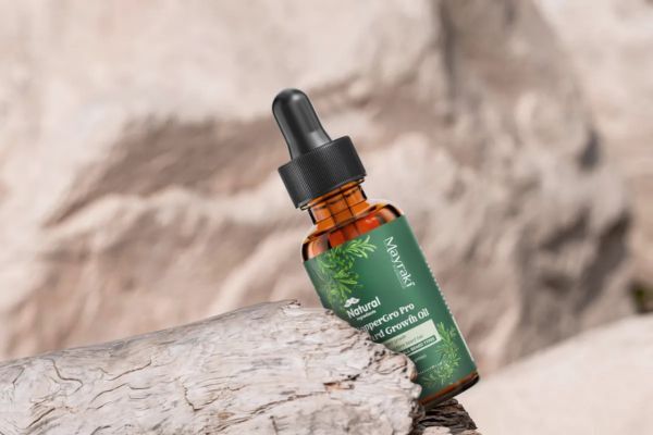 An amber dropper bottle of Mayraki Natural CopperGro Pro Beard Growth Oil rests on a textured piece of driftwood against a soft, rocky background. The natural setting emphasizes the product’s botanical and organic branding.