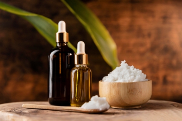 Two glass dropper bottles, one amber and one clear, sit beside a wooden bowl and spoon filled with white whipped shea butter, placed on a rustic wooden surface with green leaves in the background.