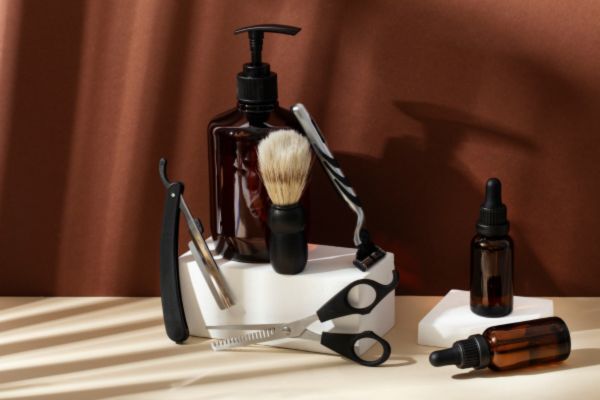A neatly arranged set of beard grooming tools including scissors, a straight razor, a shaving brush, and amber bottles with pumps and droppers, all placed on white platforms with soft shadows. The setup has a clean, minimalist aesthetic suggesting a premium grooming kit.