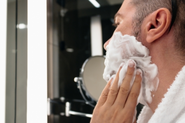 Close-up of a man applying shaving cream to his face in front of a bathroom mirror, prepping for a shave while wearing a white towel over his shoulders.