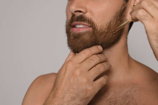 Close-up of a man applying beard oil to his thick, well-groomed beard using a dropper, highlighting one of the daily grooming habits promoted by Hair Mayraki for achieving a naturally shiny beard.