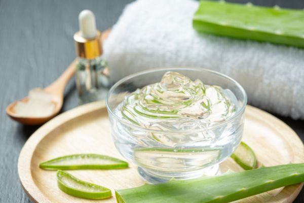 A clear glass bowl filled with aloe vera gel sits on a wooden plate surrounded by fresh aloe vera leaves, a white towel, a small glass dropper bottle, and a wooden spoon with cream.